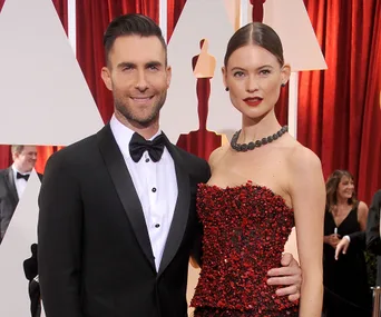 Couple in formal attire pose on a red carpet with a background of red curtains and abstract shapes.