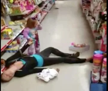 A toddler wearing pajamas stands near an unconscious woman lying on the floor of a store aisle.