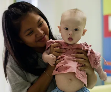 A woman holds a baby wearing a pink dress, both looking toward the camera in a bright indoor setting.