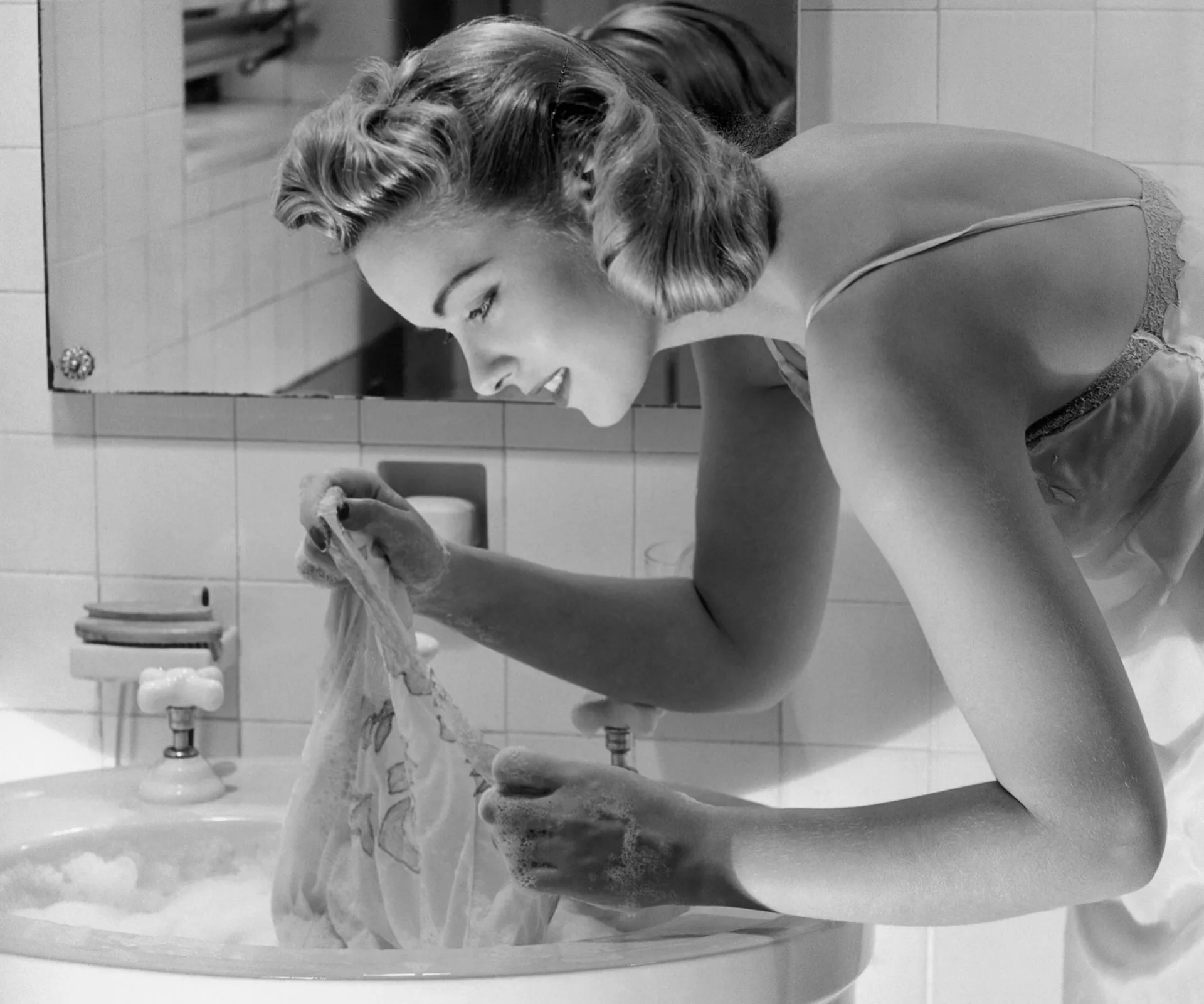 A woman in a slip hand-washes lingerie in a sink filled with soapy water, reflecting careful attention.