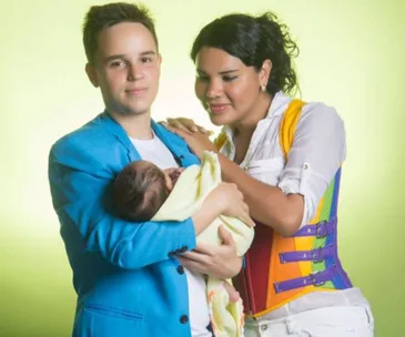 A transgender couple lovingly holds their newborn wrapped in a yellow blanket; the person on the left wears a blue jacket.