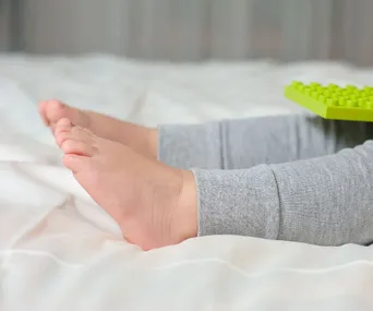 Toddler's feet in gray pants on a bed with a green toy block nearby.