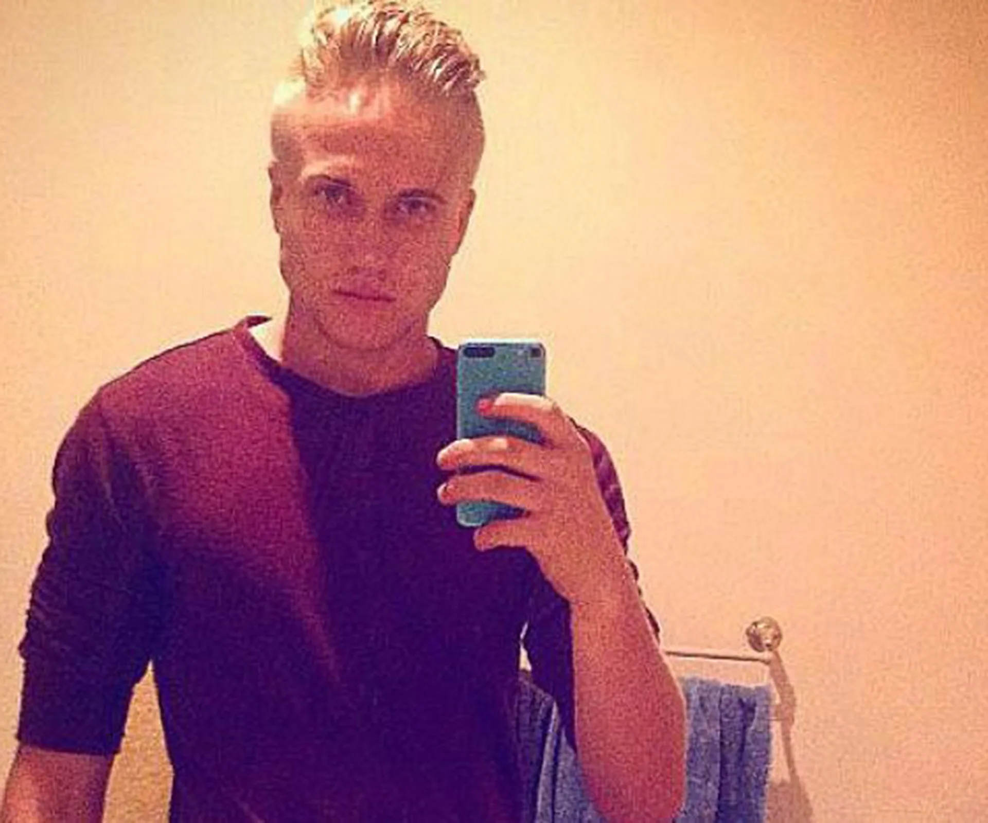 Selfie of a young man with blond hair, wearing a maroon shirt, holding a phone in front of a bathroom mirror.