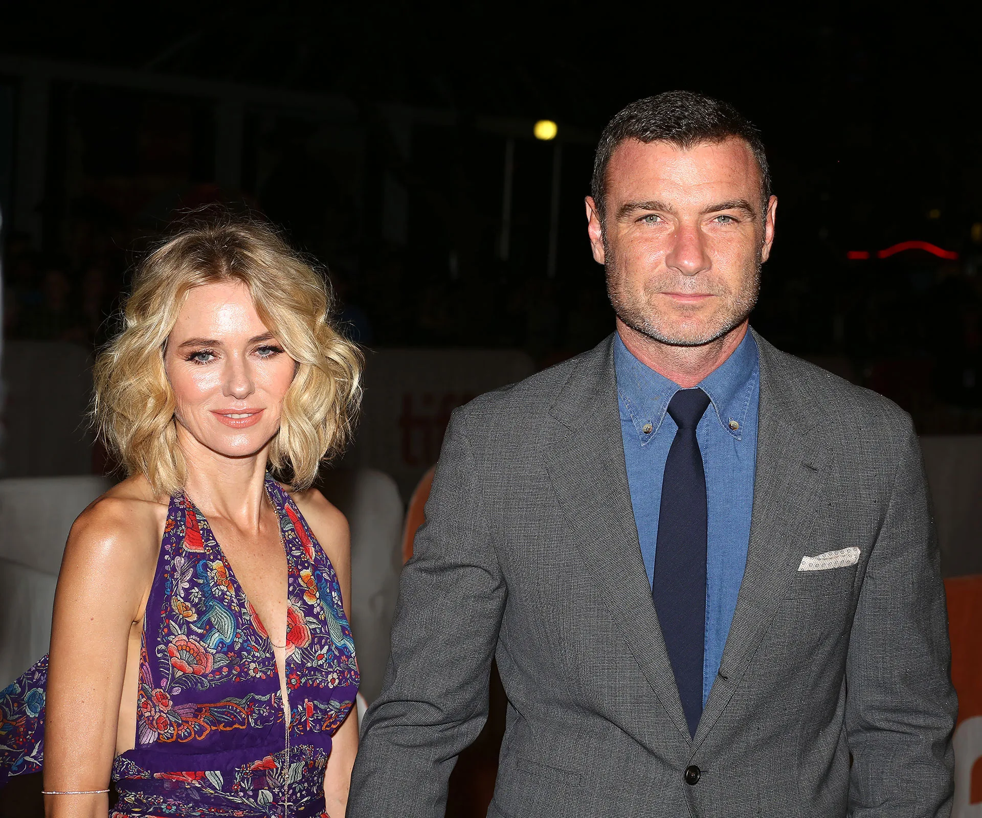 Couple posing at an event, woman in floral dress, man in gray suit and blue shirt; night-time setting.