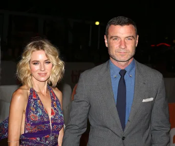 Couple posing at an event, woman in floral dress, man in gray suit and blue shirt; night-time setting.