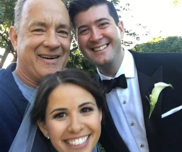 A joyful wedding selfie with a smiling man in casual attire and a happy bride and groom outdoors.