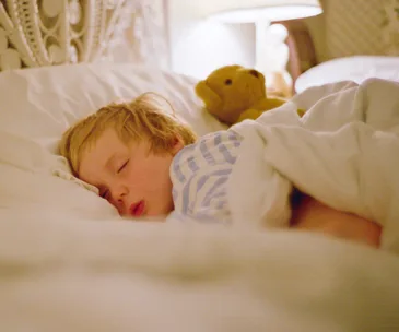 Child sleeping in bed with a teddy bear under soft lighting.