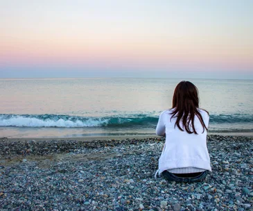 A person sits alone on a pebble beach facing the sea during a pastel-colored sunset.