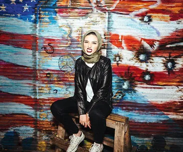 Hijabi woman in black leather jacket seated confidently in front of American flag mural.