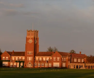 A large brick building with a clock tower and arched windows, surrounded by a grassy field at sunset.