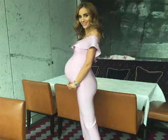 Pregnant woman in an elegant off-shoulder, pink dress standing by a dining table, smiling.