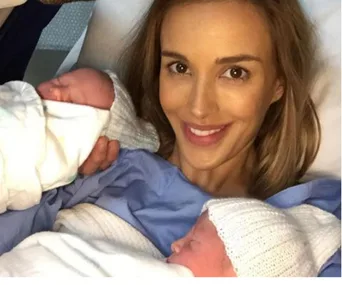 Mother in hospital bed holding newborn twins, smiling warmly.