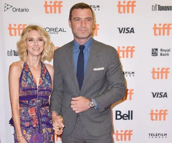Couple posing on TIFF red carpet, woman in floral dress, man in grey suit, holding hands and smiling.