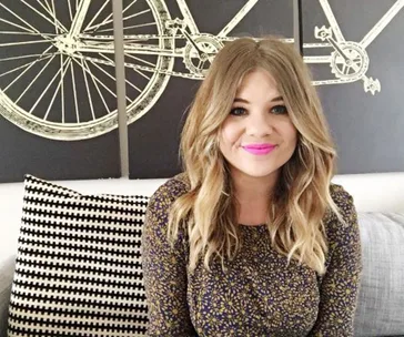 A woman with long hair and bright lipstick sits on a couch, black and white pillow, and bicycle art in the background.