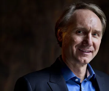 Portrait of a smiling man in a blue shirt and dark blazer against a dark background.