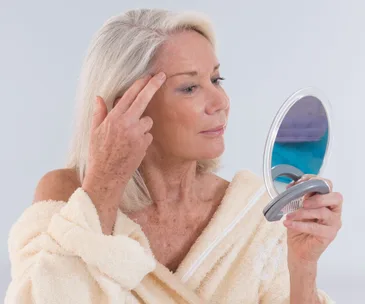 Elderly woman examines her skin in a handheld mirror, wearing a beige robe.
