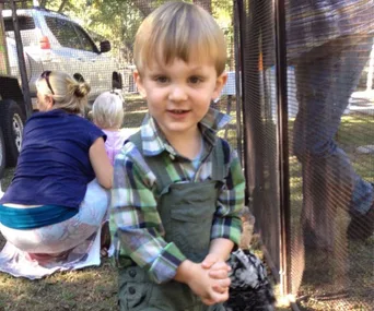 A toddler in a plaid shirt stands outdoors smiling, with adults and a vehicle in the background.