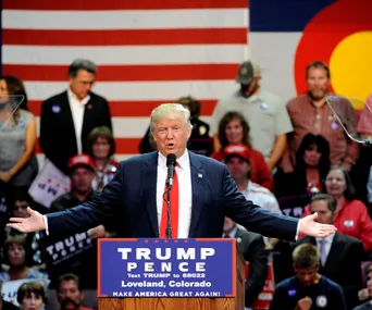A man speaks at a rally in front of an audience with a large American flag backdrop.