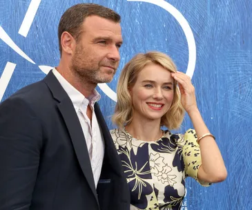 A man in a suit and a woman in a floral dress stand smiling in front of a blue patterned background.