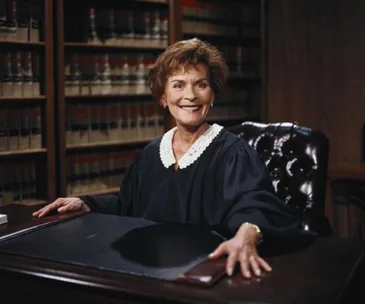 Judge in a courtroom, seated with books in the background, wearing a black robe with a white collar, smiling.