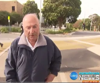 A man in a jacket walking on a path with trees and a building in the background; news logo visible.