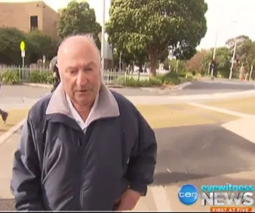 A man in a jacket walking on a path with trees and a building in the background; news logo visible.
