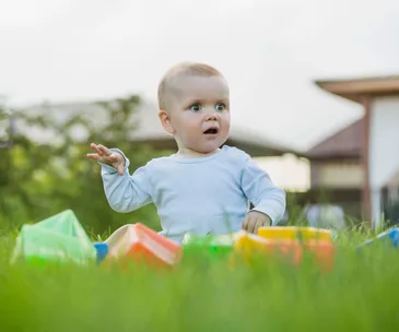 Baby sitting on grass surrounded by colorful toys, wearing a light blue shirt, looking surprised.