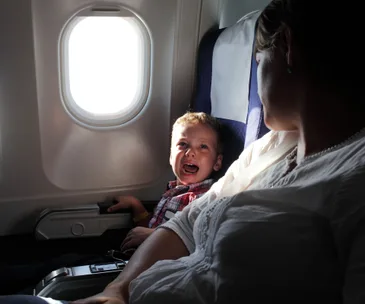 A child cries while sitting next to an adult on an airplane, near a bright window.