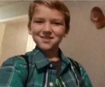 A young boy smiling, wearing a green plaid shirt, indoors with a blurred background.