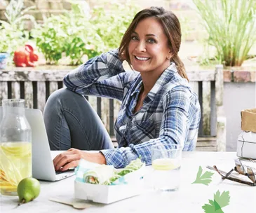 Smiling woman in plaid shirt sits at outdoor table with laptop, salad wrap, lemon water, and pear.