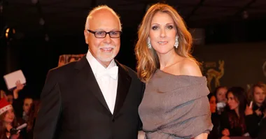 A couple poses on the red carpet, the woman in an elegant dress, and the man in a tuxedo and glasses.