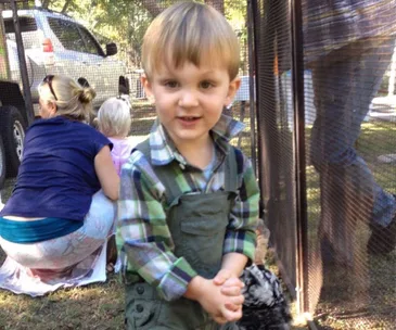 A young boy in plaid shirt stands smiling near a mesh fence; adults and a car are in the background.