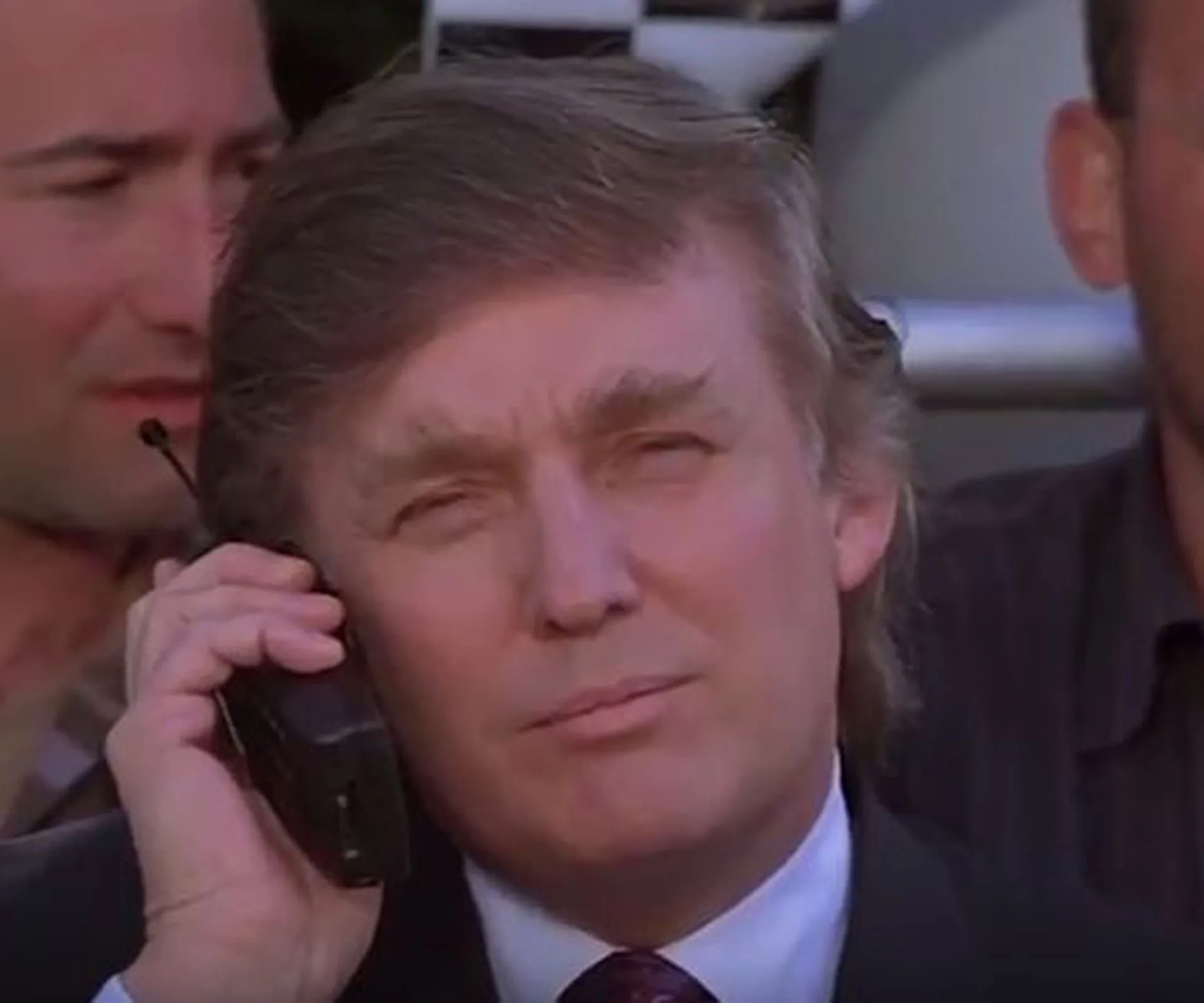 Donald Trump holding a cell phone to his ear, appearing serious, with a blurred background of people behind him.
