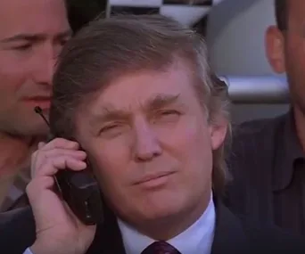 Donald Trump holding a cell phone to his ear, appearing serious, with a blurred background of people behind him.