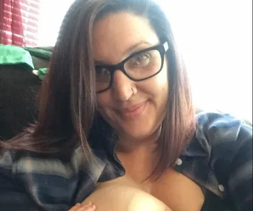 A woman with glasses breastfeeding a baby while sitting on a couch, smiling at the camera.