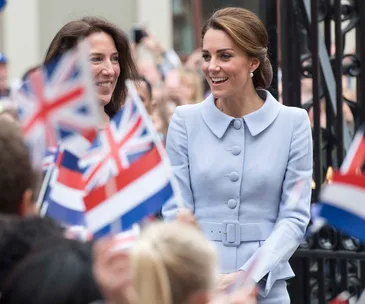 A woman in a light blue coat smiling amidst a crowd waving British flags.