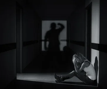 A woman sits in distress in a dark corridor with a shadow of a person raising an arm menacingly on the wall.