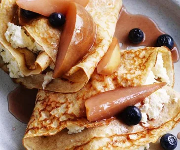 Crepes with ricotta, poached pears, blueberries, and syrup on a plate.