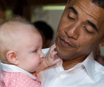 A man gently holds a baby in a pink outfit, who is touching his face, both sharing a tender moment.