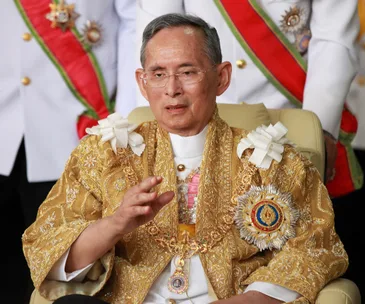 Elderly man in ornate gold robe with medals, gesturing, seated, with others in decorative uniforms in the background.