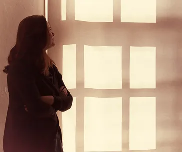 A woman stands facing windowpanes, sunlight casting soft grid-like shadows on the wall.
