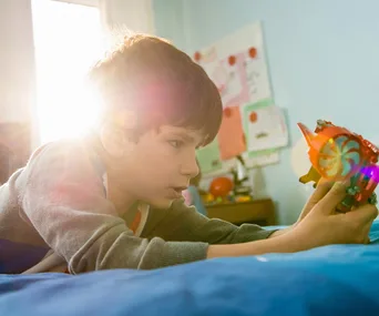 Young boy playing with a toy on a bed, sunlight streaming from a window behind him.