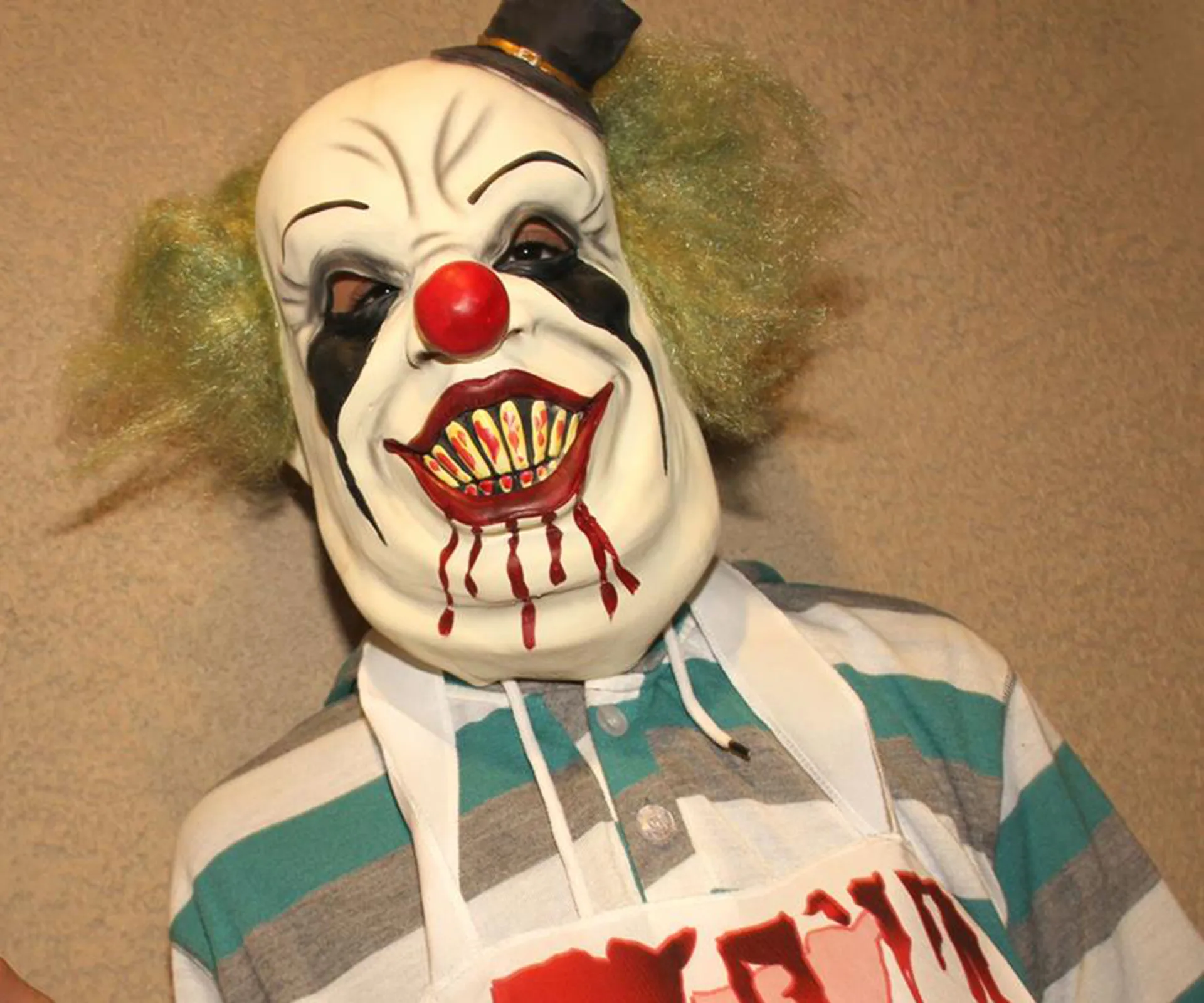 Creepy clown mask with green hair, red nose, and sharp teeth, wearing a striped shirt and apron with red lettering.