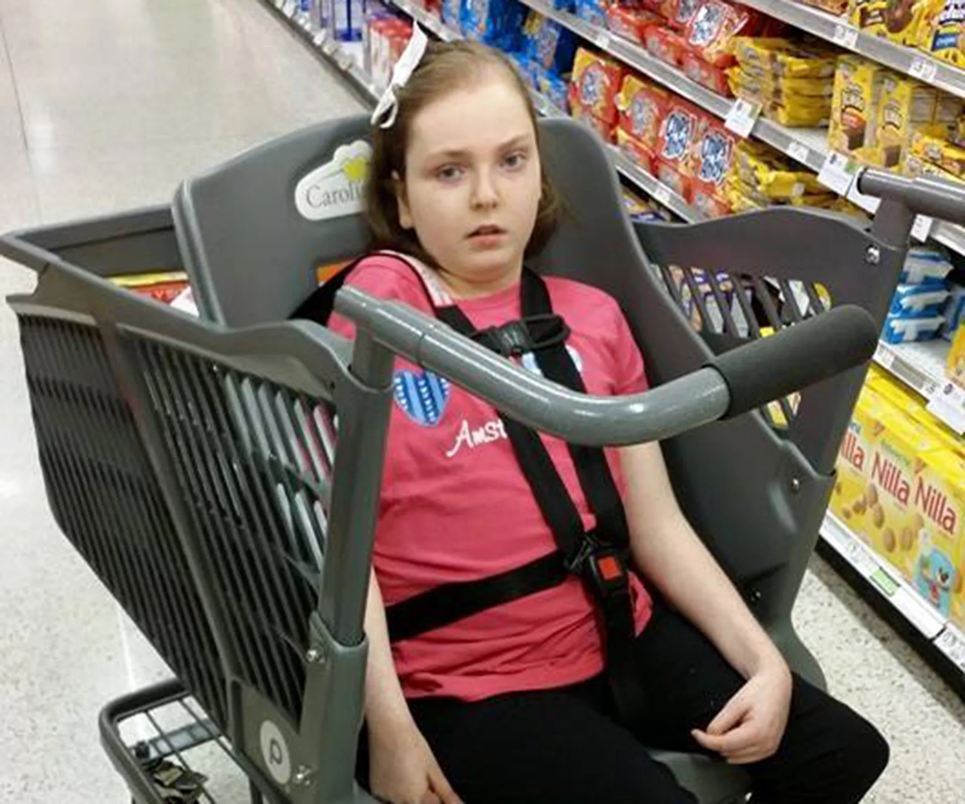 Child seated in Caroline's Cart in a grocery store aisle, wearing a pink shirt and black pants.