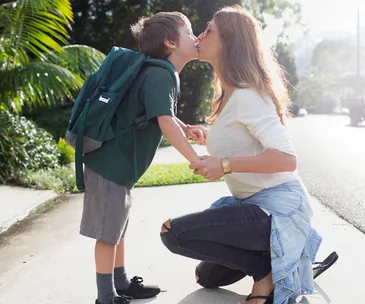Mother kneels to kiss her young son wearing a school backpack on a sunny sidewalk.