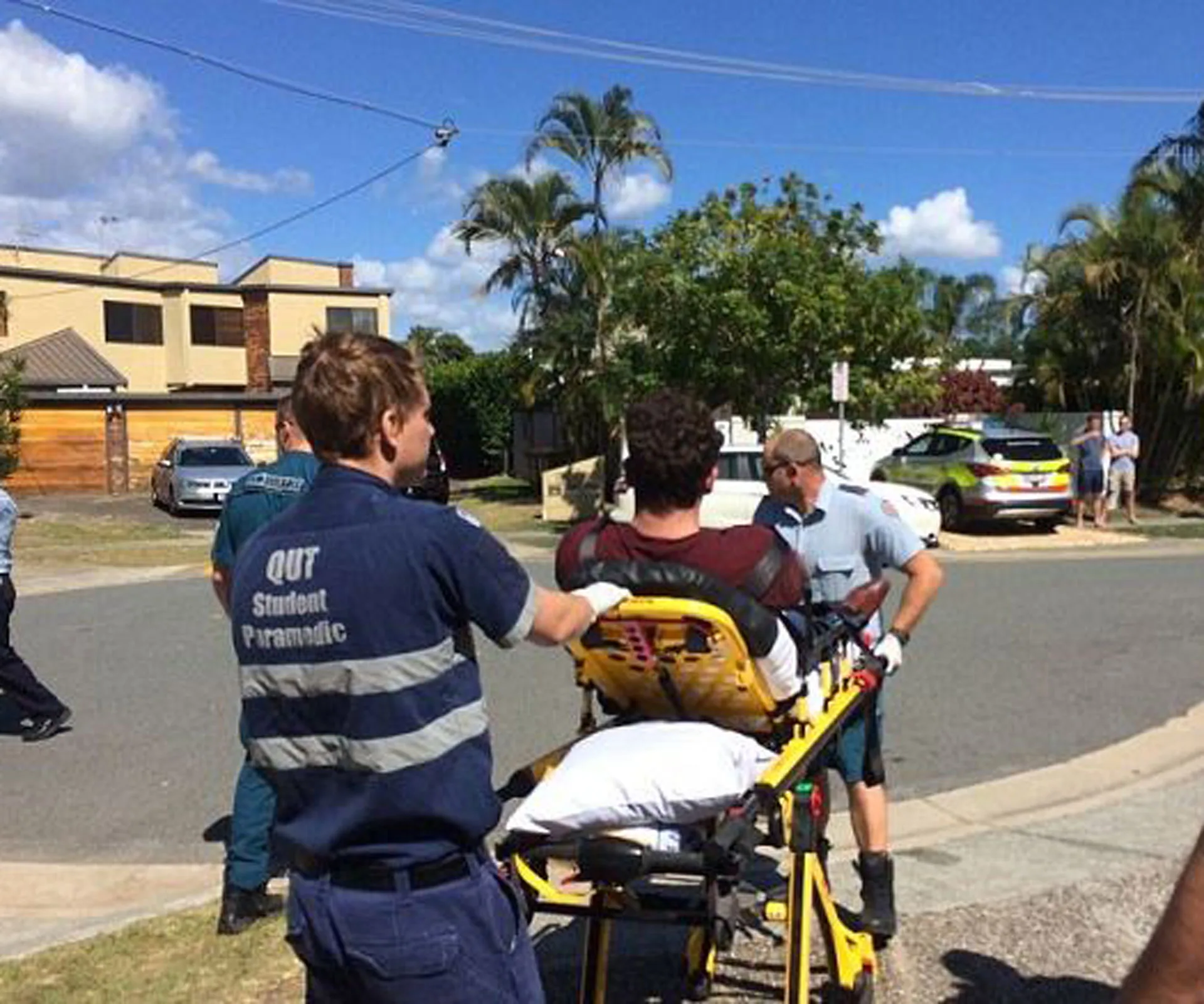 Paramedics assisting a person on a stretcher in a suburban street, with emergency vehicles and bystanders nearby.