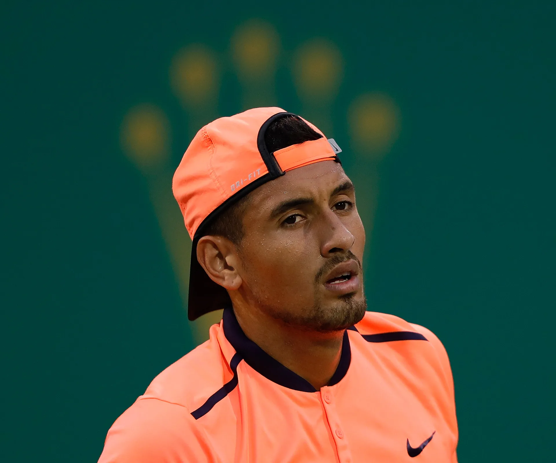 Tennis player in an orange outfit and cap facing forward, green background with blurred logo.