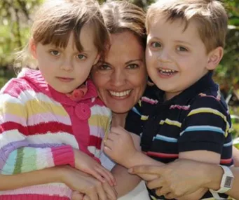 A mother embraces her young daughter and son, all smiling, dressed in colorful striped sweaters, in a garden setting.