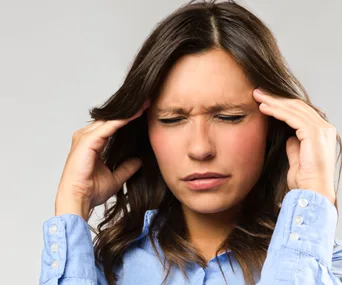A woman in a blue shirt clutching her temples, appearing to suffer from a headache or migraine.