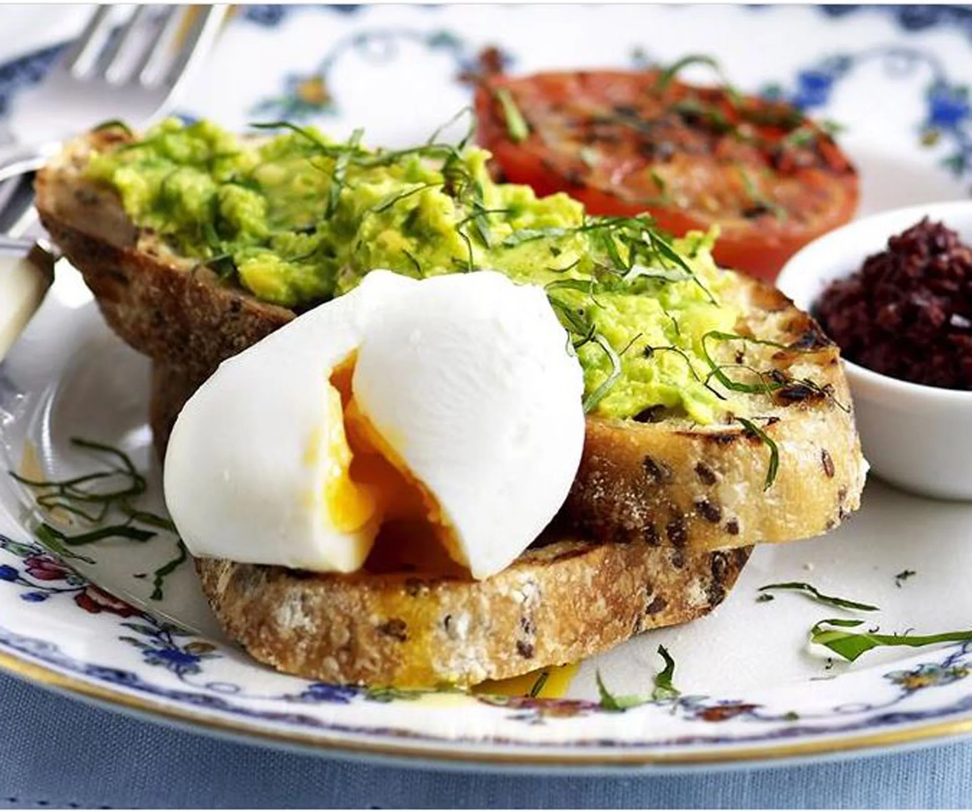 Plate with smashed avocado on toast, poached egg, grilled tomato, and a small dish of relish, garnished with herbs.